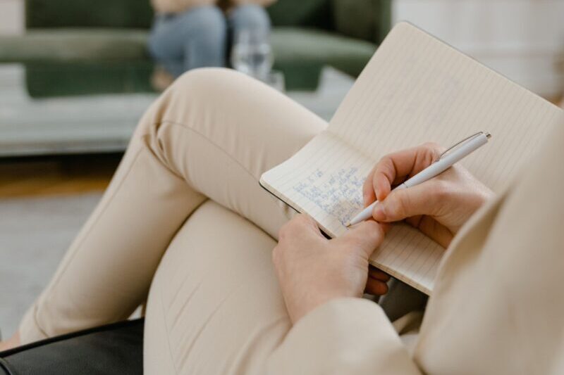 A therapist takes notes in a modern office setting during a therapy session with a patient.